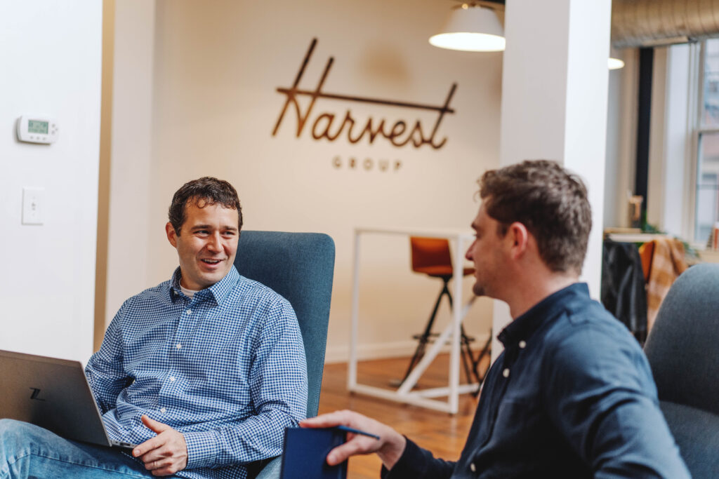 two men sitting in office talking with "Harvest Group" logo on the wall in the background