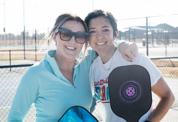 two women, one wearing sunglasses, standing together with pickleball paddles after playing outside at a company event.